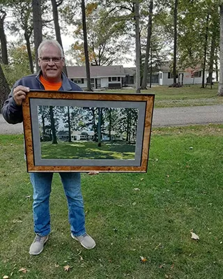 Customer holding a framed custom landscape paint by numbers featuring a cherished home surrounded by tall trees