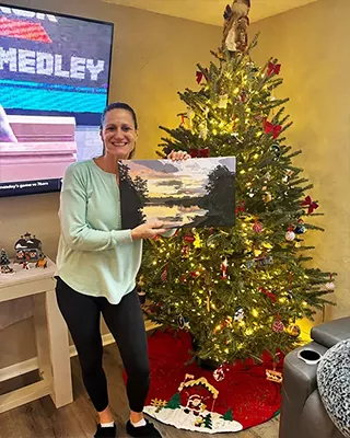 Woman holding a finished custom paint by numbers landscape beside a Christmas tree